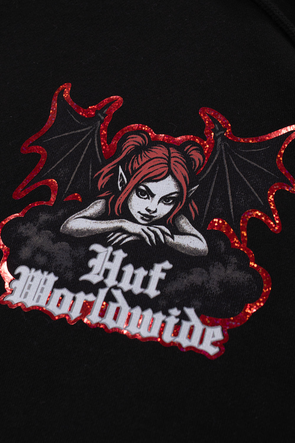 Close-up detailed view of the screen-printed chest graphic on the black HUF Chaos Hoodie, showing the devilish red-haired anime-style succubus character, dark smoke clouds, Gothic white "Huf Worldwide" typography, and the textured metallic red glitter foil border running along the perimeter of the artwork