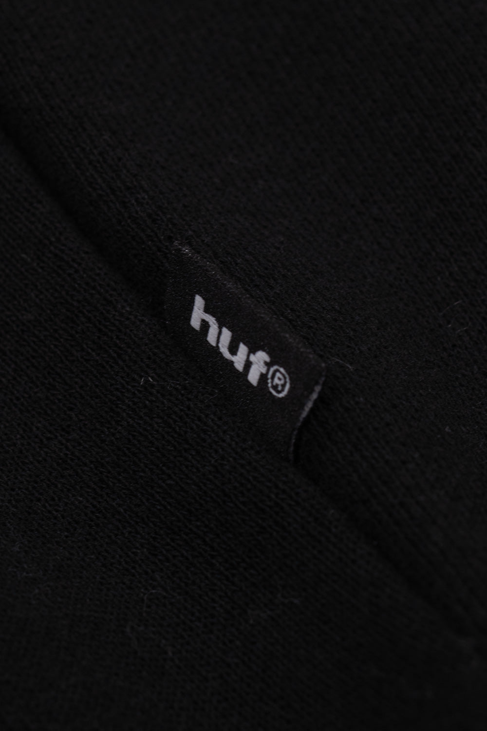 Macro shot of the subtle black woven HUF brand flag label with white registered logo lettering, neatly stitched into the side seam near the lower hem of the heavyweight black hooded fleece sweatshirt