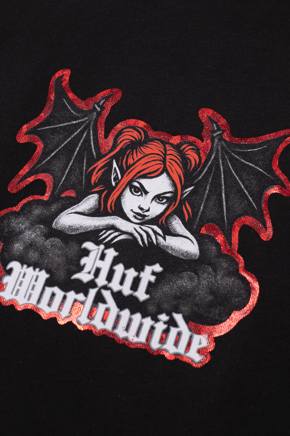 Macro close-up shot of the front chest graphic on the black HUF Chaos T-Shirt, displaying the detailed screen-printed horned succubus character with red hair, bat wings outlined in metallic red glitter, dark smoke clouds, and the textured white Gothic "Huf Worldwide" branding across the black cotton fabric