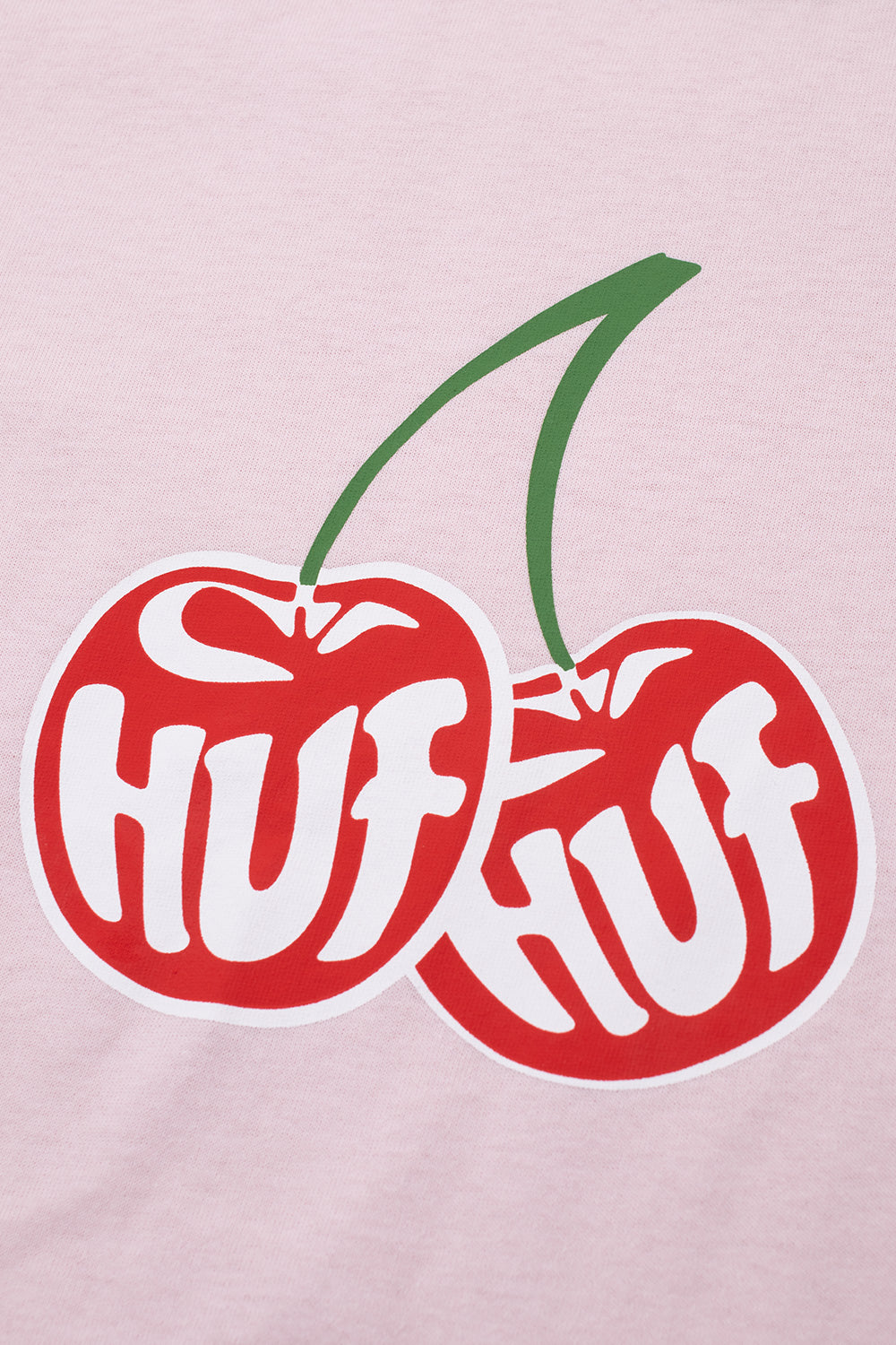 Detailed close-up shot of the front chest graphic on the dusty pink HUF Cherries Ringer T-Shirt, highlighting the vibrant red paired cherries with green stems and white stylized bubble text reading "HUF" on each fruit, set against the pastel pink cotton weave.