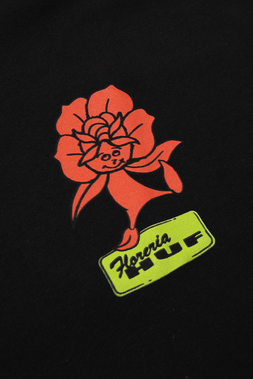 Macro close-up shot of the left chest print on the black HUF Flower District T-Shirt, highlighting the smiling orange cartoon rose bloom figure balanced on a bright lime-green logo badge with script "Floreria" text and bold black "HUF" block lettering