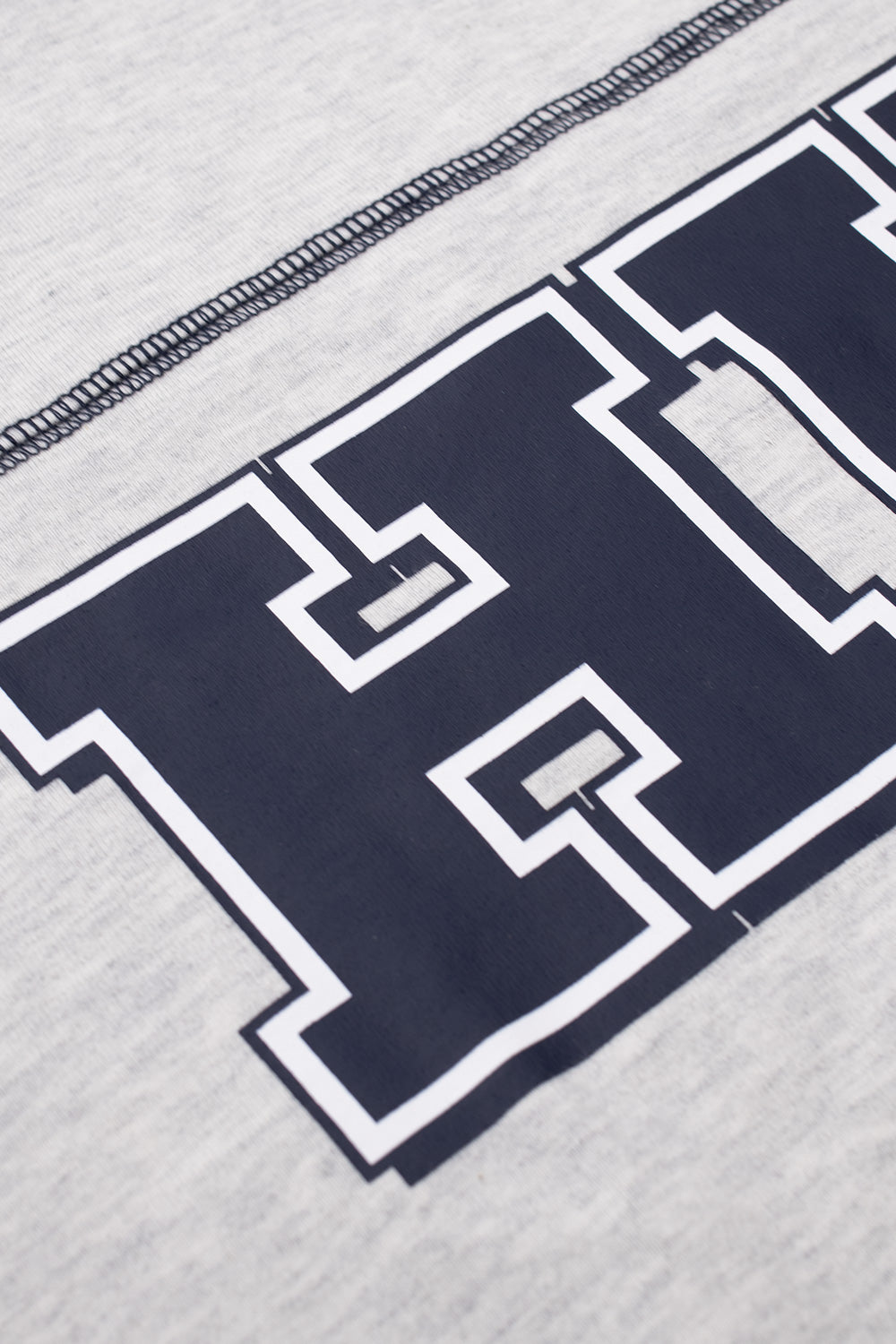 Detailed close-up shot of the front chest graphic on the heather grey HUF Legacy Football tee, showcasing the navy blue varsity collegiate HUF lettering with double white perimeter borders beneath exposed navy overlock seam stitching.