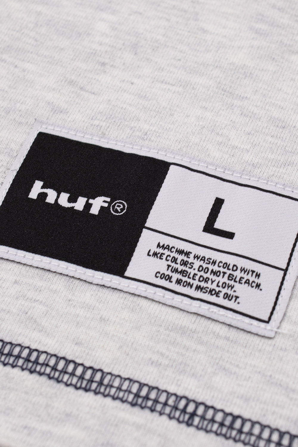 Macro shot of the black and white woven athletic jock tag label stitched above the bottom hem of the heather grey HUF shirt, displaying the HUF brand name, size L indication, and printed garment care instructions.