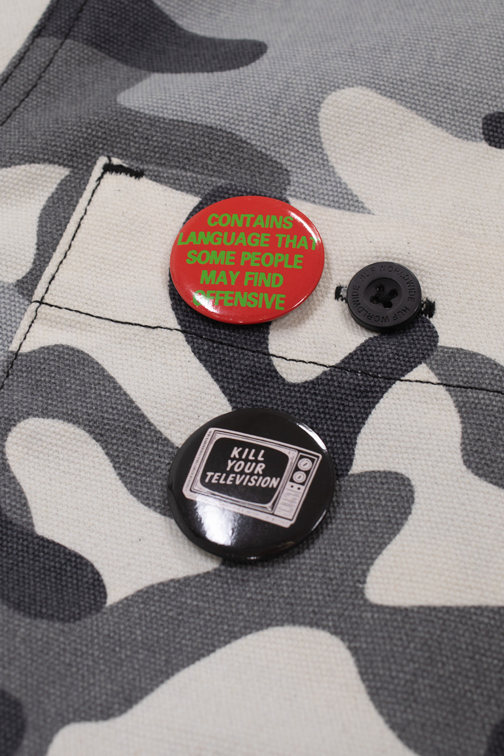 Macro detail shot of the right chest pocket on the urban camo duck canvas jacket, showing an engraved black HUF Worldwide button alongside a red button pin reading Contains Language That Some People May Find Offensive and a black Kill Your Television graphic pin badge.