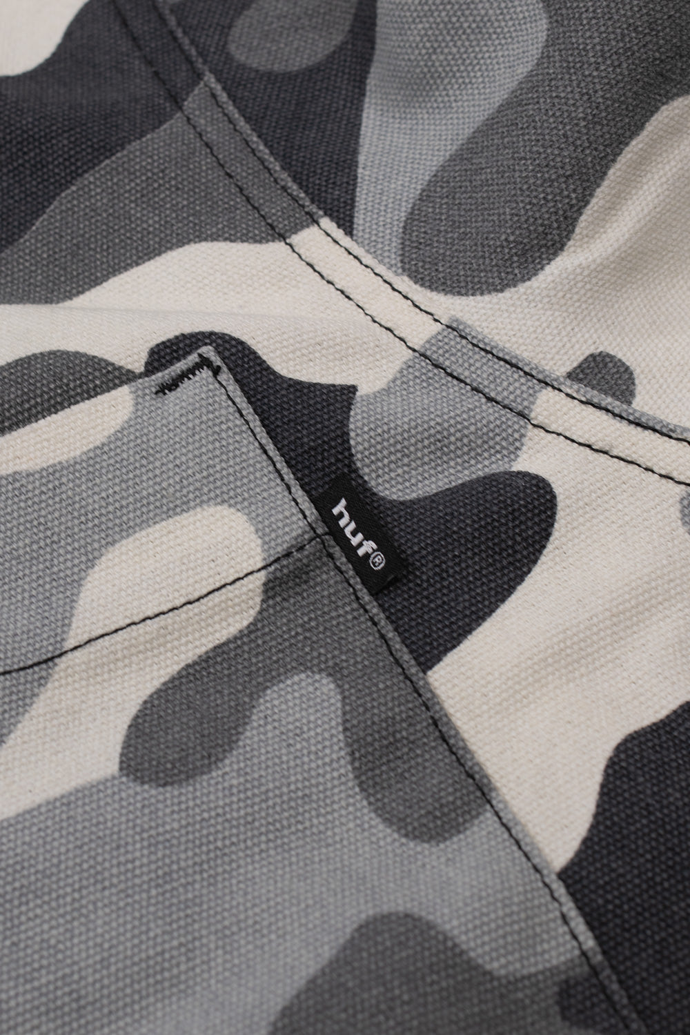 Detailed view of the left chest flap pocket on the HUF Olympia Camo shirt jacket, highlighting the heavy-textured 324g duck canvas weave in grey and black camouflage and the black woven HUF logo flag label sewn into the side seam.
