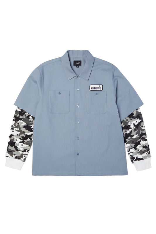Front flat lay view of the HUF Shadows Long Sleeve Work Shirt in light blue, featuring a cotton-viscose blend short-sleeve work shirt body with spread collar, full button-front closure, two buttoned chest flap pockets, a black-and-white embroidered HUF star patch on the left chest, and contrast sewn-in long sleeves in grey and white star-camo with white ribbed knit cuffs