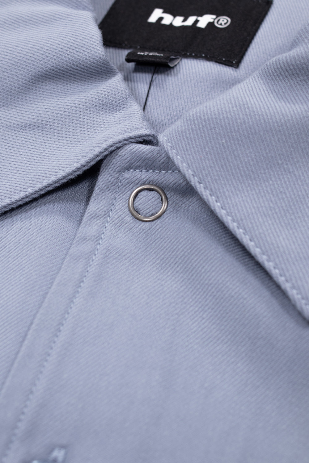 Macro close-up shot of the collar and upper placket on the light blue HUF Shadows Work Shirt, showing the structured twill weave, fine seam stitching, top metallic eyelet button, and the woven black "HUF" neck tag