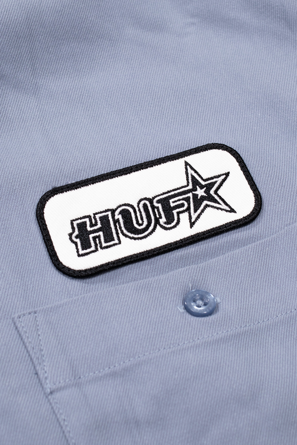 Detailed close-up of the left chest pocket on the HUF Shadows Work Shirt, highlighting the light blue translucent button, double-stitched pocket hem, and the sewn-on rounded rectangular patch featuring black embroidered "HUF" lettering with a shooting star icon on a white background with a black merrowed border