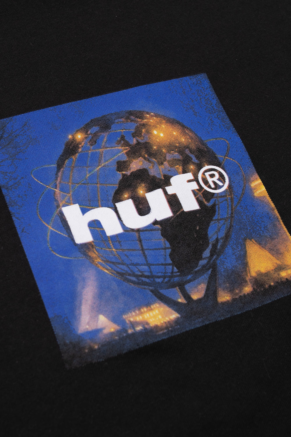 Detailed angled close-up shot of the center chest graphic on the black HUF Unisphere tee, displaying the illuminated blue and orange night photography of the Flushing Meadows Unisphere steel globe structure contrasted against bold white HUF typography and deep black cotton knit fabric.