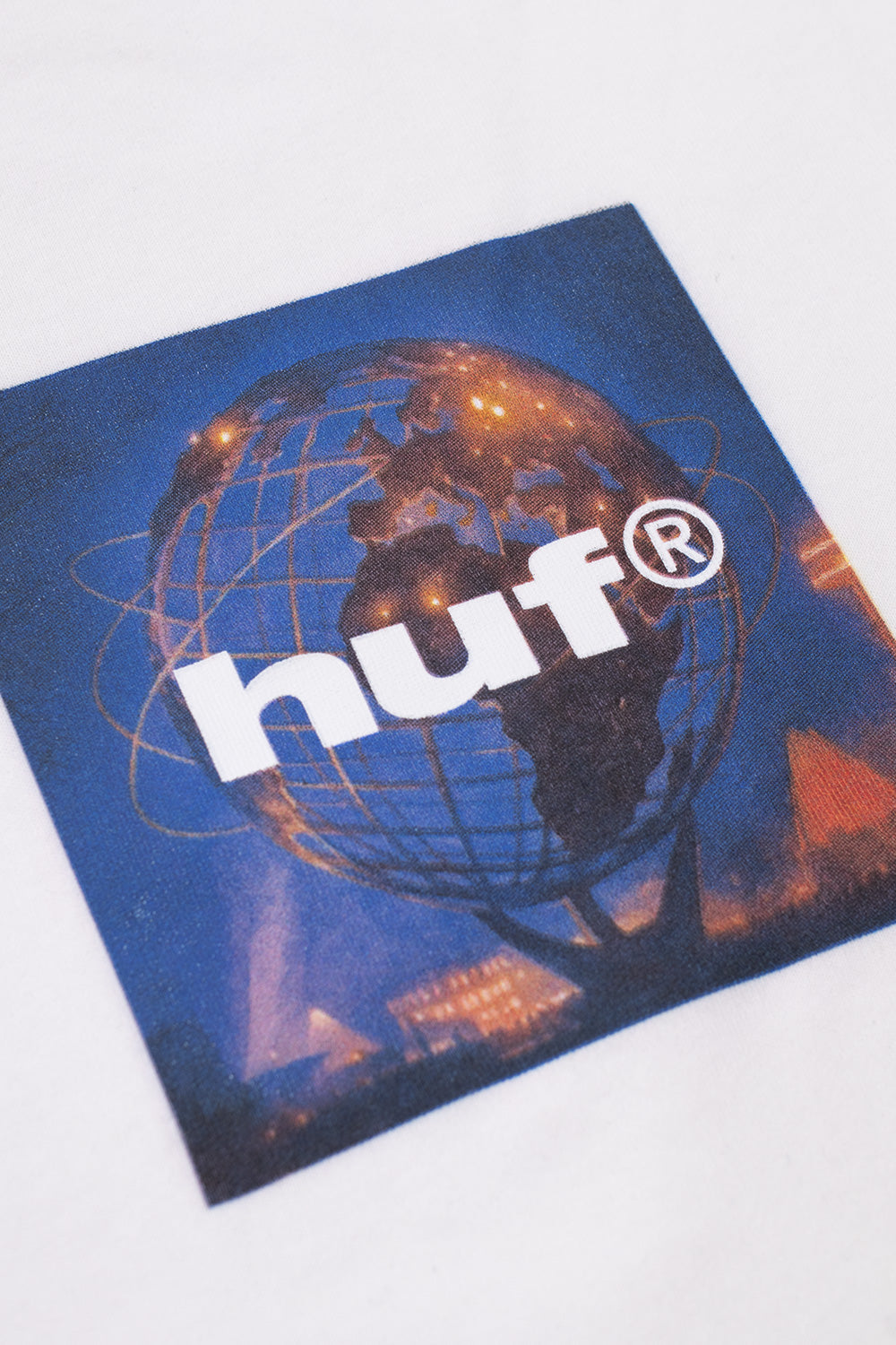 Detailed angled close-up shot of the center chest graphic on the white HUF Unisphere tee, highlighting the photographic night shot of the glowing blue and orange Flushing Meadows Unisphere globe structure beneath clean white screen-printed HUF lettering.