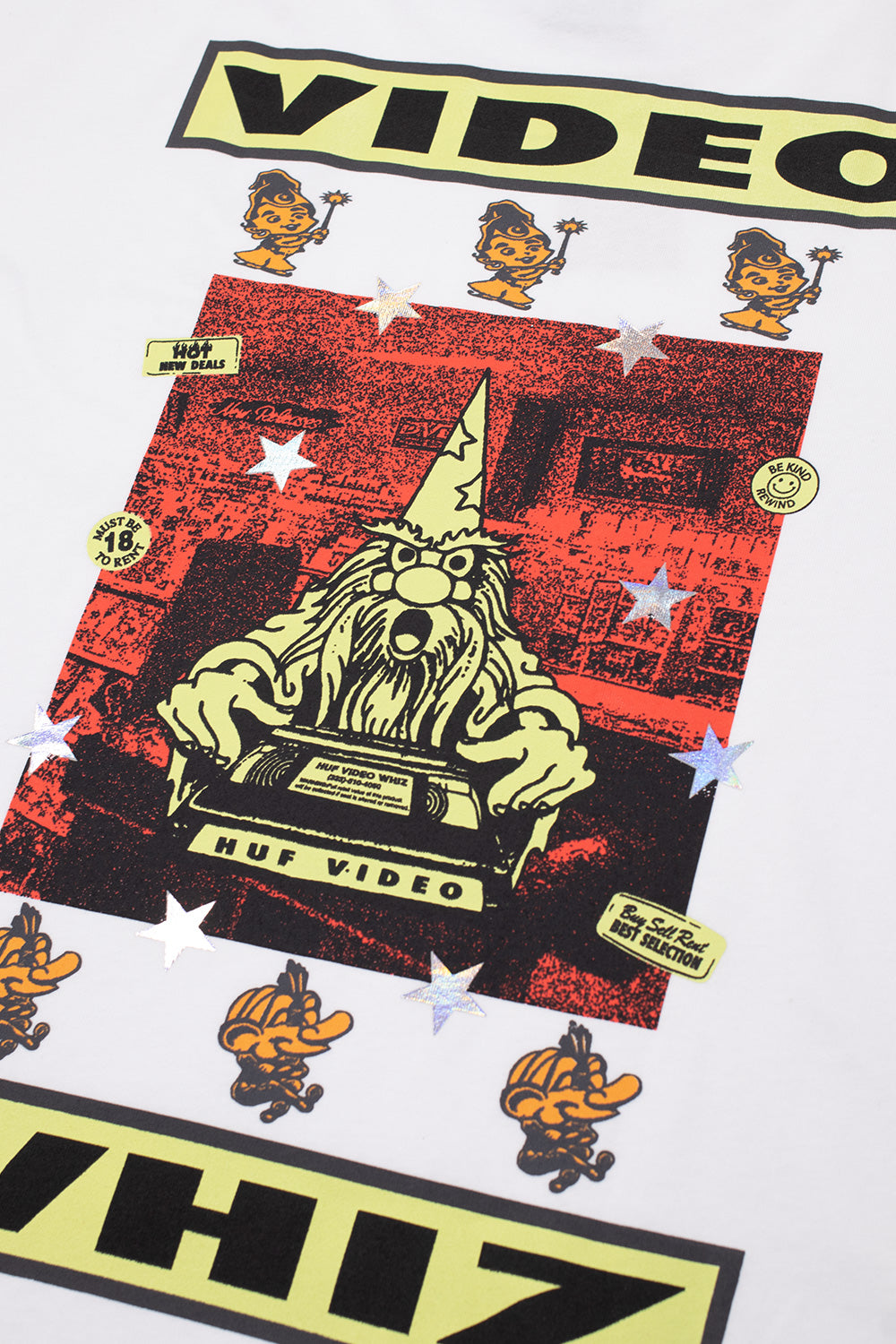 Detailed close-up shot of the back graphic print on the white HUF Video Genie T-Shirt, highlighting the wizard character holding a branded "HUF VIDEO" cassette against a red video store backdrop with holographic stars, "Be Kind Rewind" badges, and small smiling genie mascots