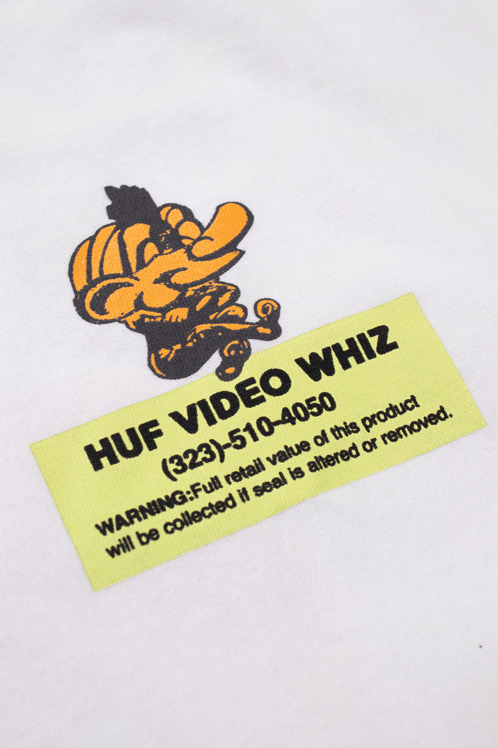 Macro close-up shot of the left chest graphic on the white HUF Video Genie T-Shirt, displaying the textured screen print of a yellow turban-wearing genie character above a lime-yellow rental label reading "HUF VIDEO WHIZ" with a phone number and warning notice across the white cotton weave