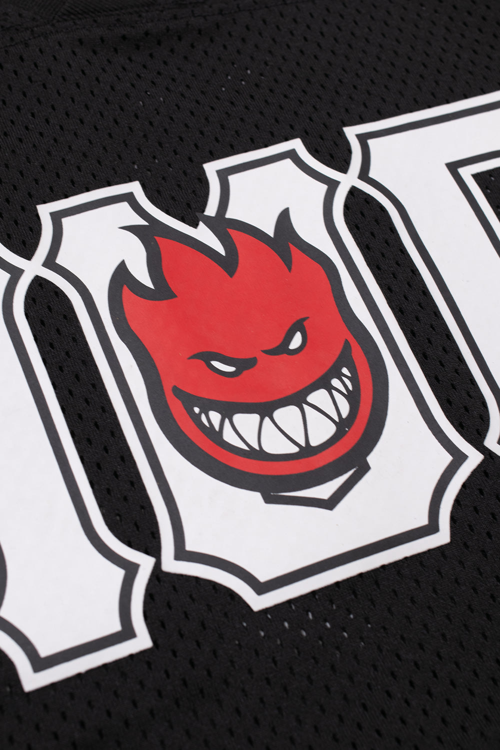 Detailed close-up shot of the front chest graphic on the HUF X Spitfire jersey, highlighting the red and black Spitfire Wheels Bighead character with sharp grinning teeth framed inside the white collegiate letter U between the white-bordered HUF typography on black athletic mesh.