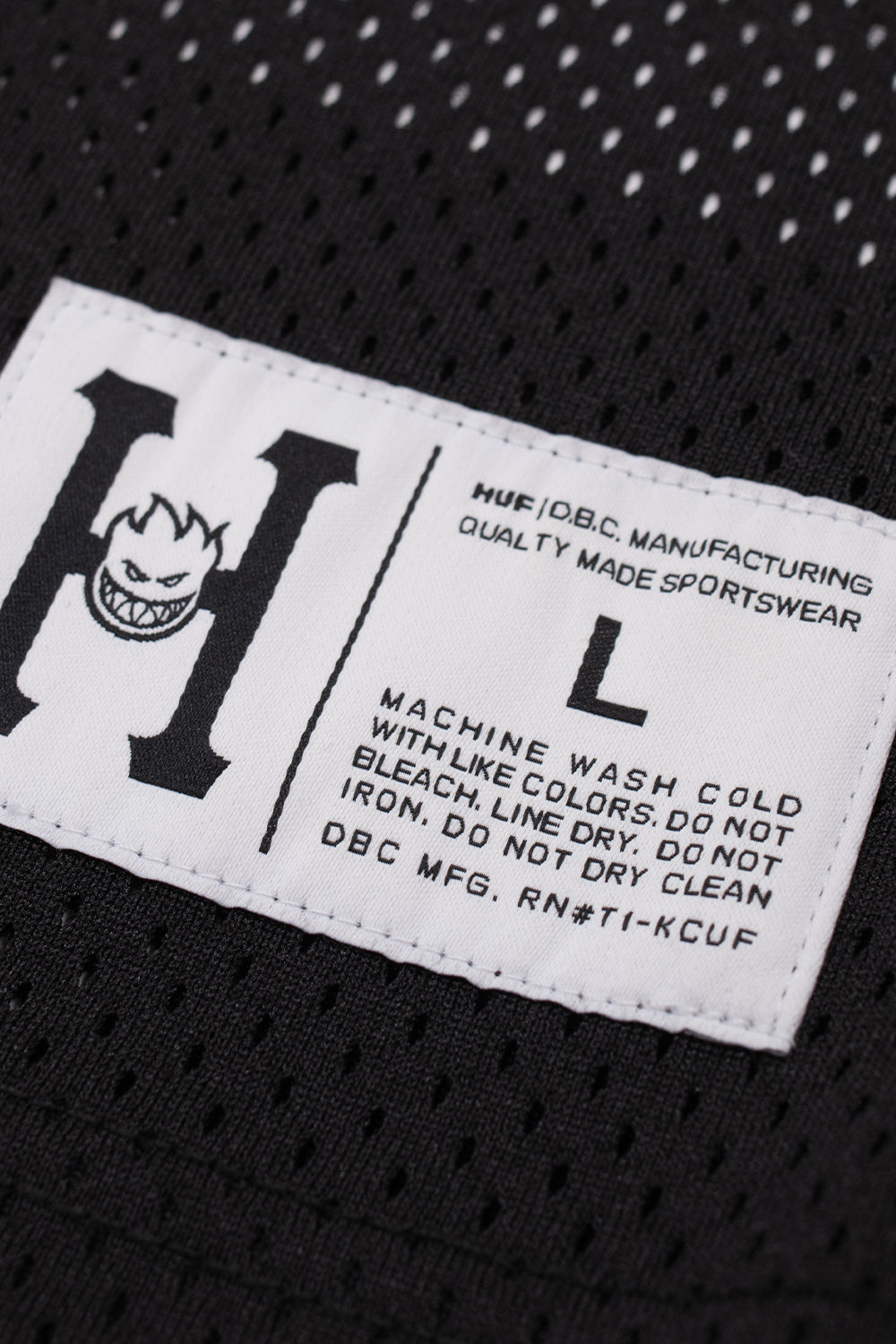 Close-up view of the collaborative white woven athletic locker patch stitched to the lower front hem of the jersey, featuring a black collegiate H containing the Spitfire Bighead logo, size L label, HUF D.B.C. Manufacturing branding, and garment care text.