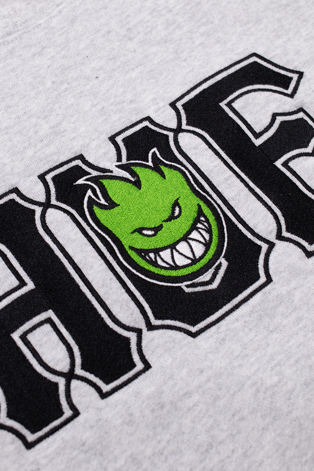 Detailed close-up shot of the high-density co-branded chest embroidery on the heather grey HUF X Spitfire pullover, showing the black outlined HUF text and the vibrant green Spitfire Bighead character with a grinning toothy face.