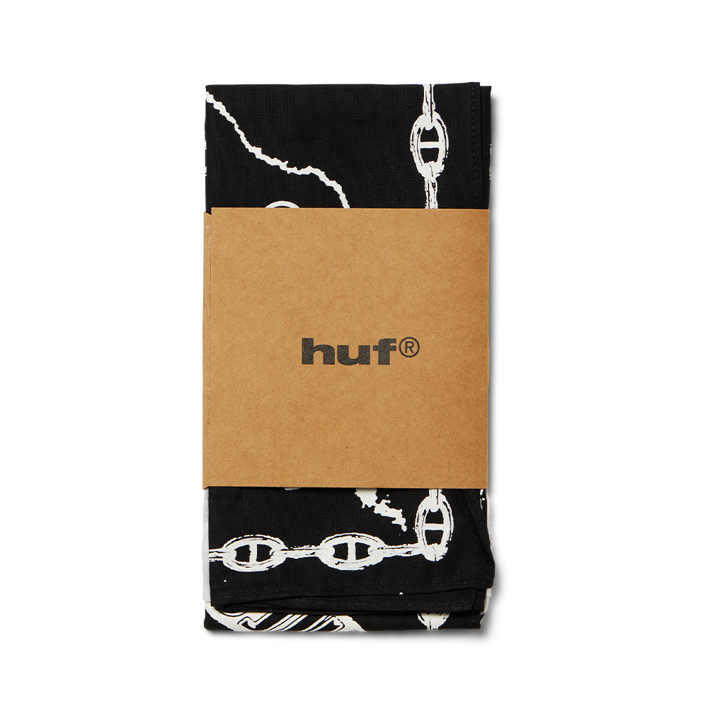 Neatly folded studio presentation of the black Huf X Three 6 Mafia Hypnotize Minds Bandana, wrapped in a natural brown cardboard packaging sleeve featuring the centered black printed lowercase "huf®" logo, displaying the chain border print and lightning accents.