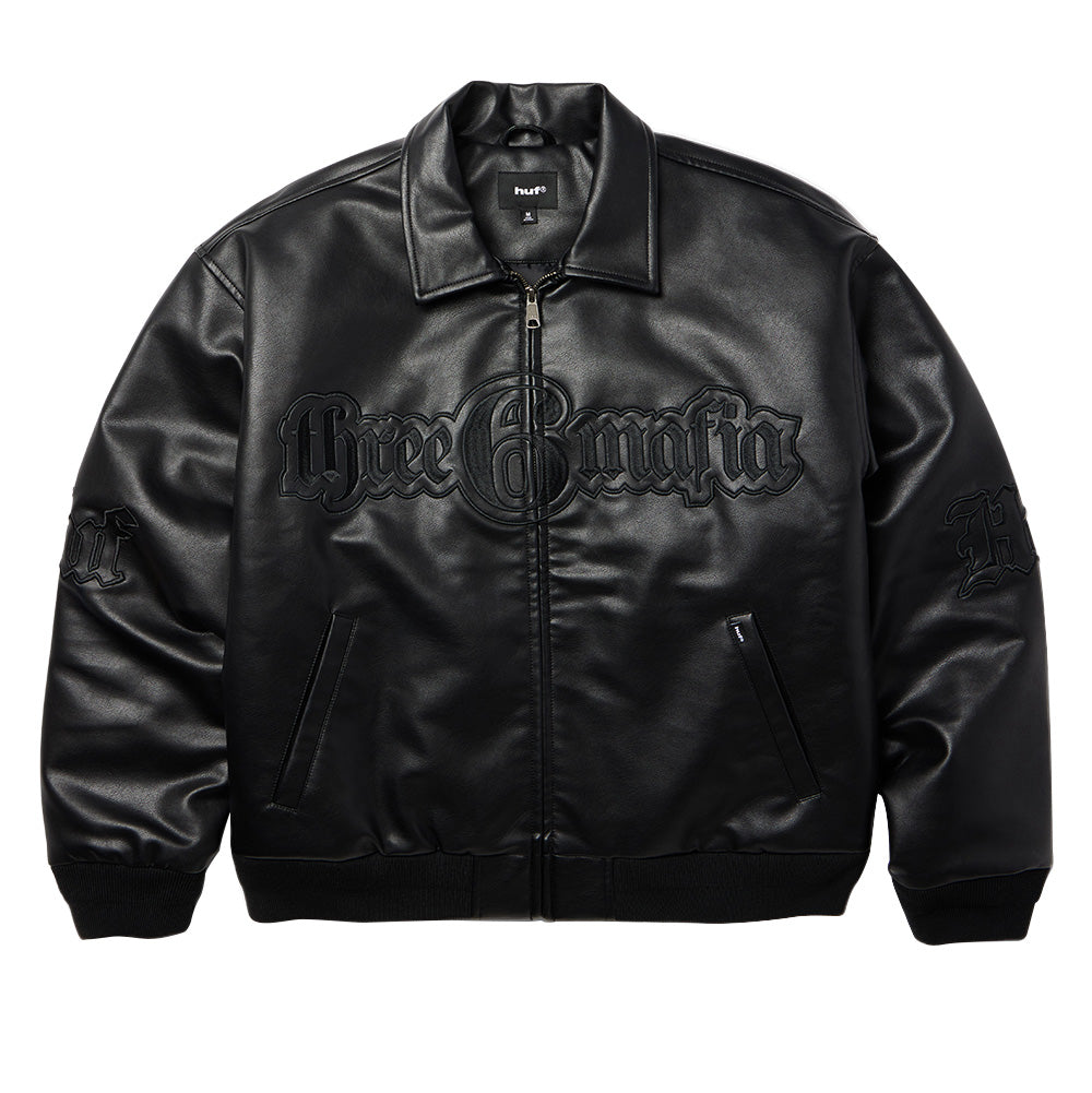 Front flat lay view of the black Huf X Three 6 Mafia jacket, showing the grained faux leather construction, fold-down collar, silver metal zipper closure, welt hand pockets, ribbed cuffs and hem, and a large tonal black embroidered Three 6 Mafia circular emblem across the chest.