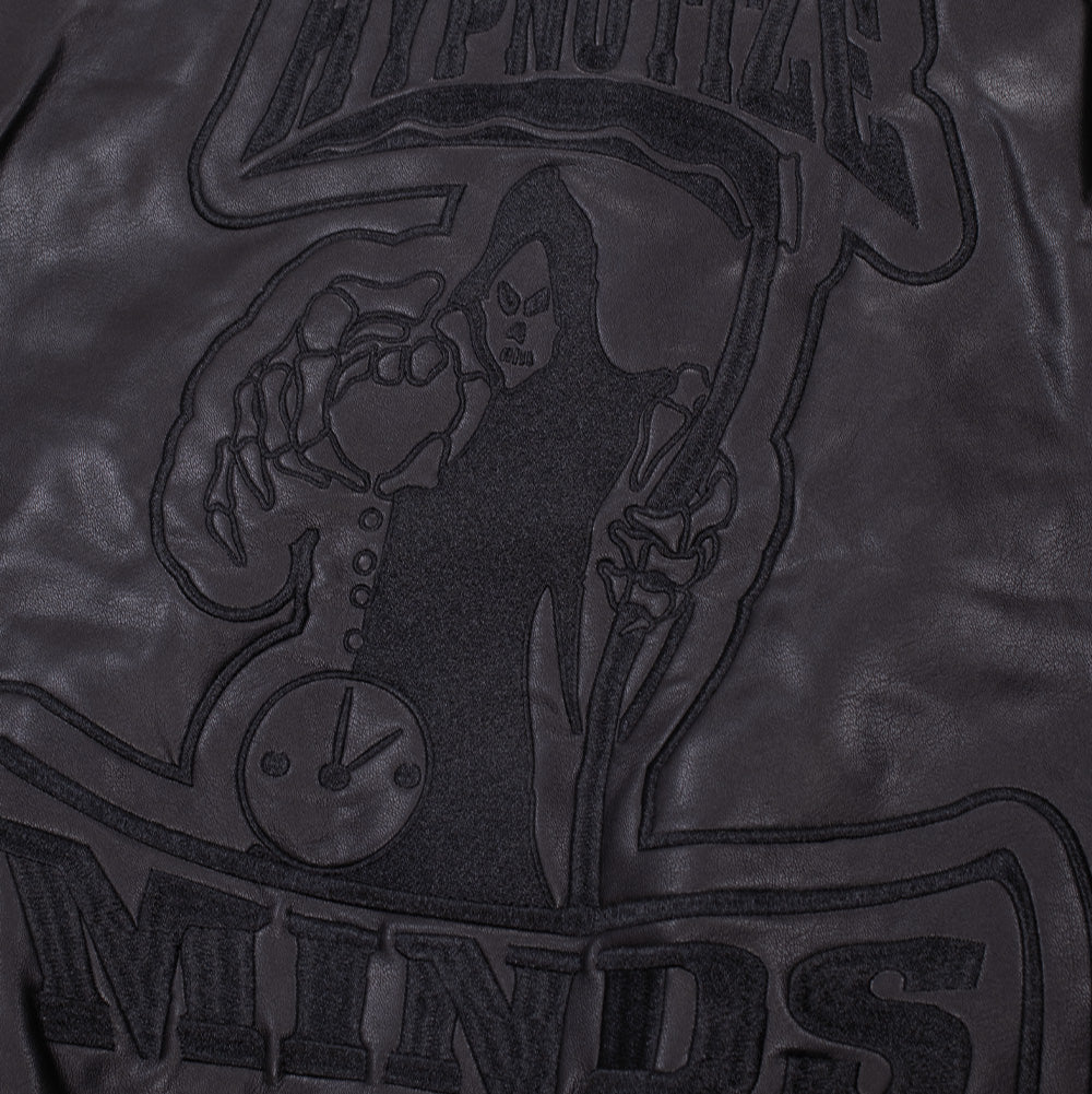 Detailed macro studio close-up of the back panel on the Huf X Three 6 Mafia jacket, highlighting the tonal black satin-stitched embroidery and raised faux leather patch displaying the Hypnotize Minds reaper, hourglass clock face, and arched lettering.