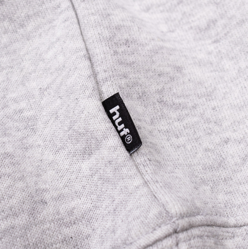 Studio macro detail of the lower side seam on the Huf X Three 6 Mafia Memphis Hoodie, focusing on the small black woven HUF brand flag label with clean white embroidery set against the thick heather grey fleece and rib-knit waistband seam.