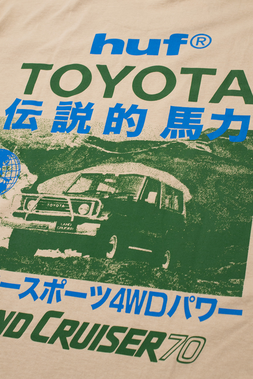 Detailed macro close-up of the back graphic on the sand Huf X Toyota Land Cruiser 70 SS T-Shirt, displaying the screen-printed green halftone photographic scene of the FJ70 4x4 off-road vehicle with bull bar, bright blue globe icon, and dynamic Japanese Katakana typography on the heavyweight cotton fabric.