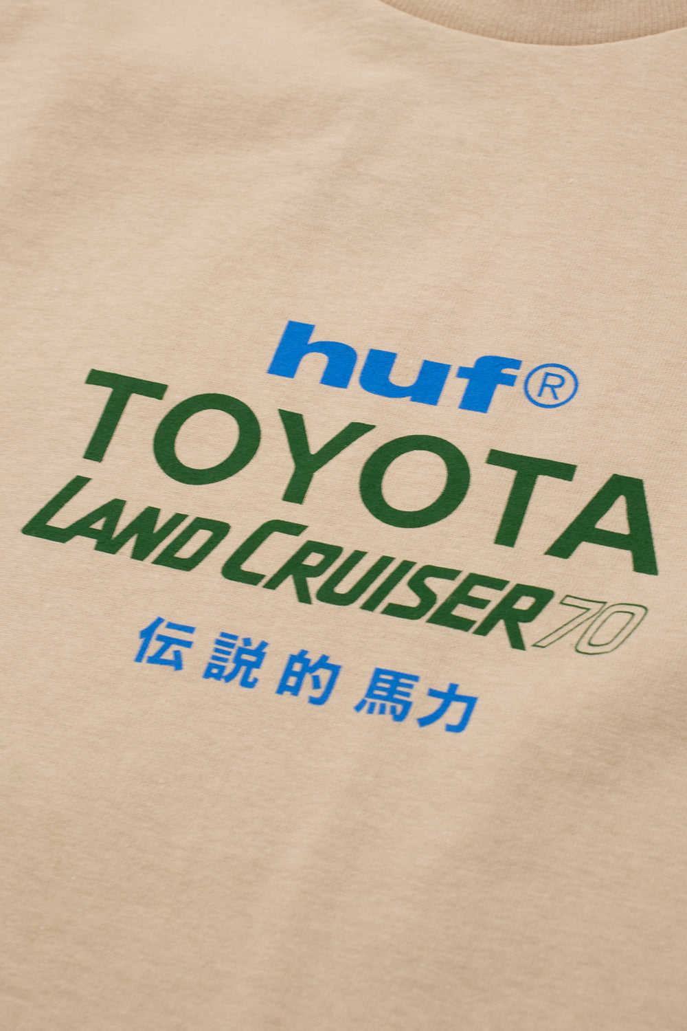 Macro studio close-up of the center front chest graphic on the Huf X Toyota Land Cruiser 70 SS T-Shirt, highlighting the clean screen print of the blue lowercase "huf®" logo above dark green "TOYOTA", green outlined "LAND CRUISER 70", and blue Kanji characters on the textured sand-colored cotton jersey.