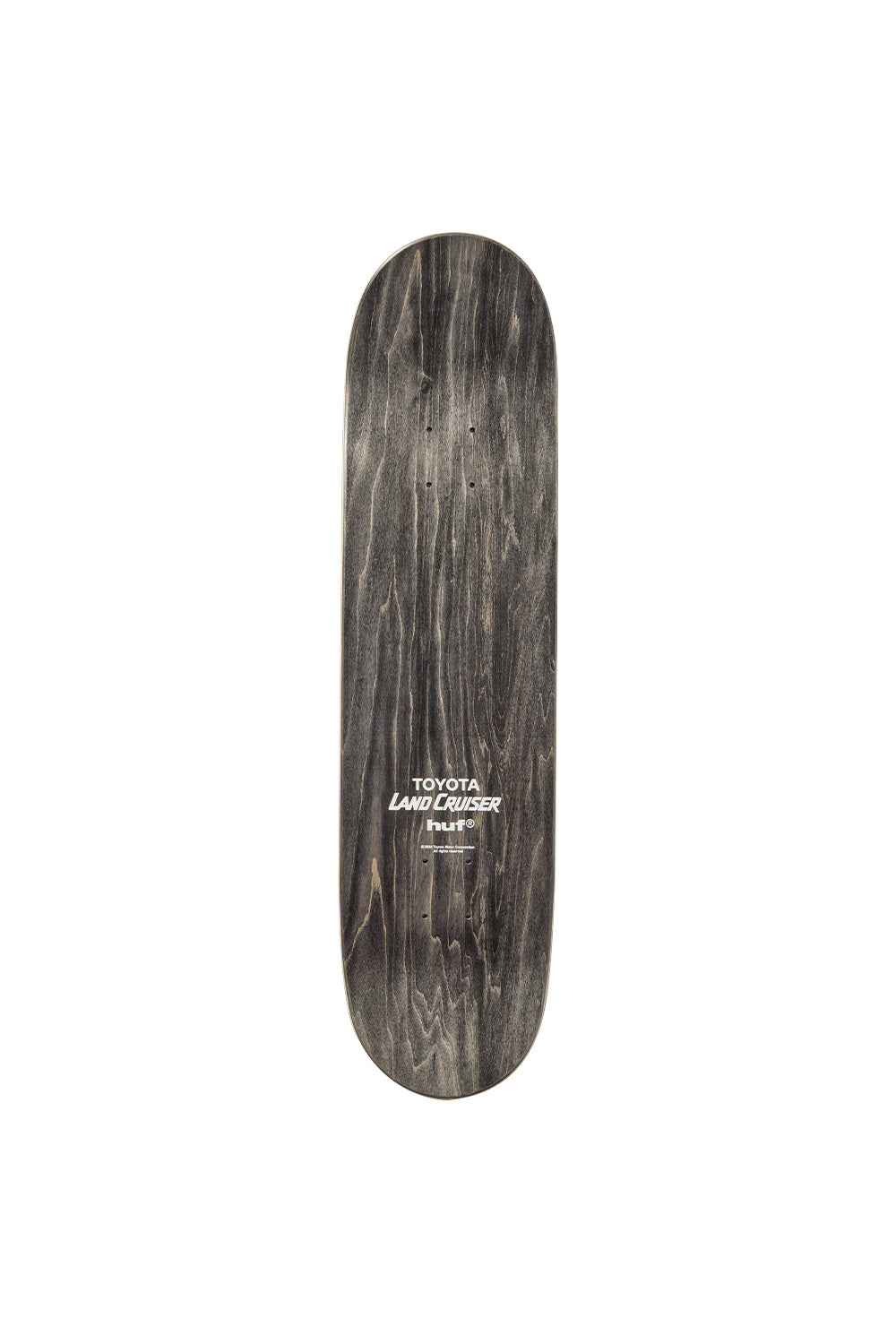 Top side view of the Huf X Toyota Land Cruiser skateboard deck, showing a dark charcoal stained wood grain finish on the premium 7-ply Canadian maple construction, complete with truck mounting drill holes and a centered white screen-printed collaboration graphic featuring stacked "TOYOTA", italicized "LAND CRUISER", and "huf®" brand logos.