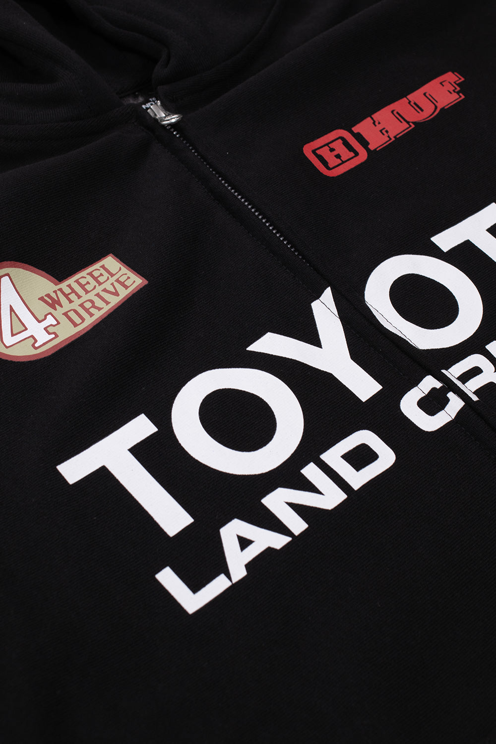 Detailed macro close-up of the upper chest graphics on the black Huf X Toyota Land Cruiser Heavyweight FZ Hoodie, showing the bold white screen-printed "TOYOTA LAND CRUISER" typography split by the zipper, alongside the vintage cream-and-red "4 WHEEL DRIVE" crest and red outlined "HUF" block logo.