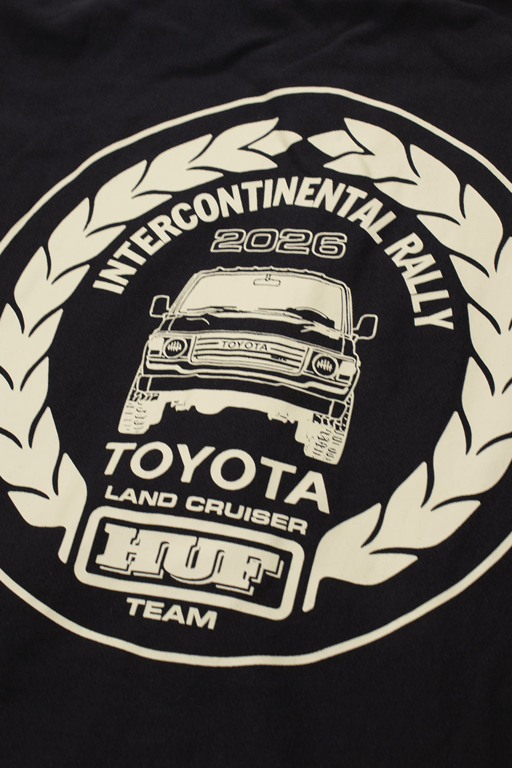 Studio macro shot of the back graphic on the Huf X Toyota Land Cruiser Heavyweight FZ Hoodie, highlighting the cream off-white screen-printed rally crest with laurel branches, the detailed line art of the off-road vehicle with bull bar, and bold motorsport event lettering on the dense black fleece fabric.