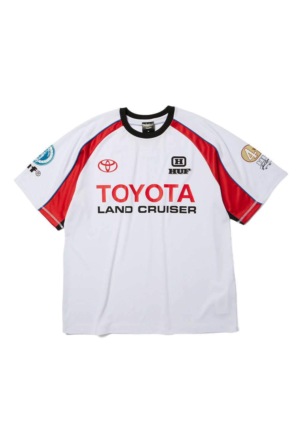 Front flat lay view of the Huf X Toyota Land Cruiser Rally Jersey in white, featuring a relaxed-fit short-sleeve racing silhouette made from 180-gram polyester knit fabric with a black ribbed crewneck collar, red raglan sleeve inserts with blue piping, a red Toyota oval emblem, black-and-white HUF badge, bold red "TOYOTA" and black "LAND CRUISER" screen prints across the chest, and printed rally decals on both sleeves.