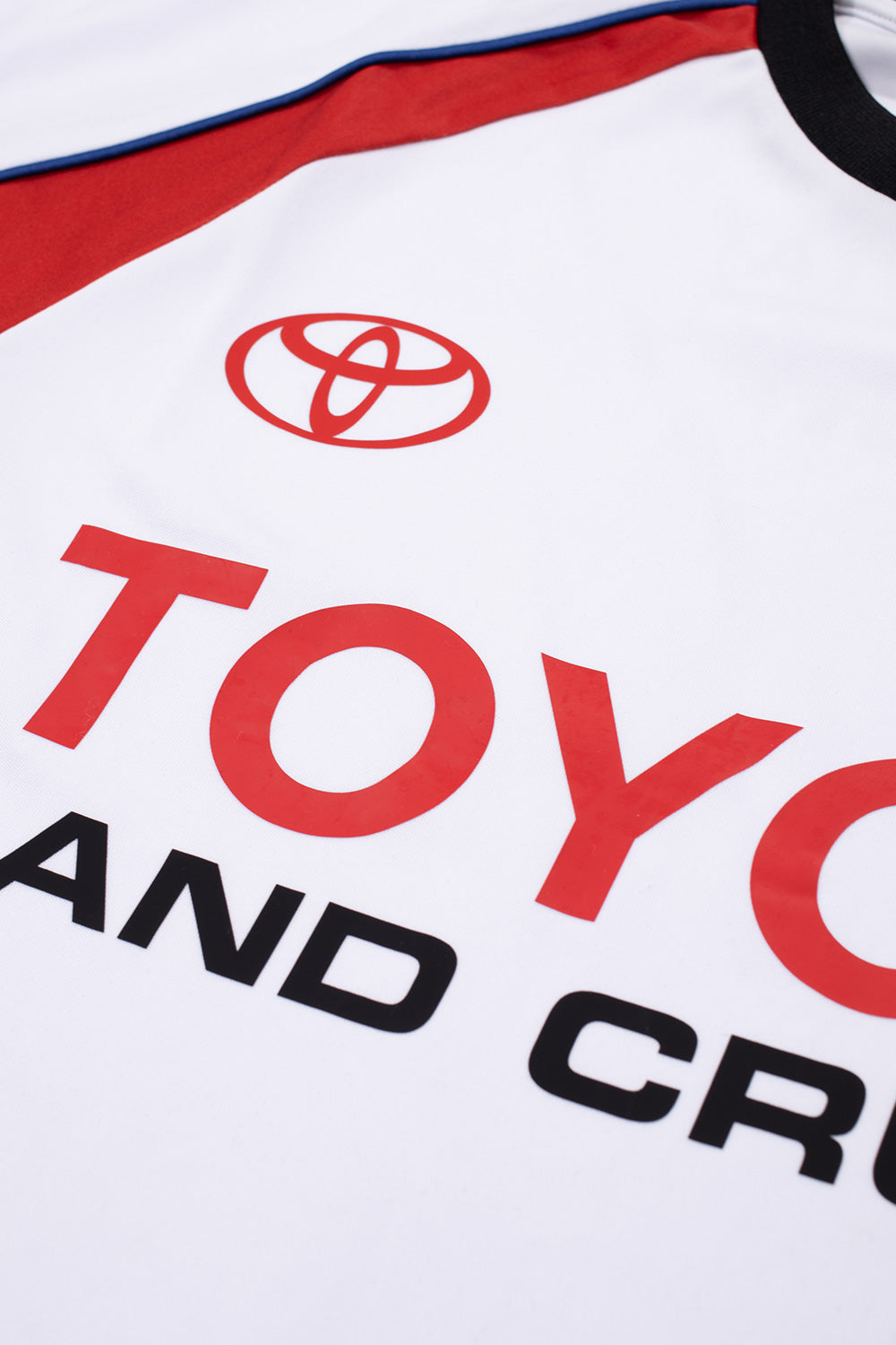 Macro close-up shot of the center chest print on the Huf X Toyota Land Cruiser Rally Jersey, highlighting the crisp red Toyota corporate logo emblem and bold capital lettering of "TOYOTA" alongside black "LAND CRUISER" typography on the white breathable polyester fabric.