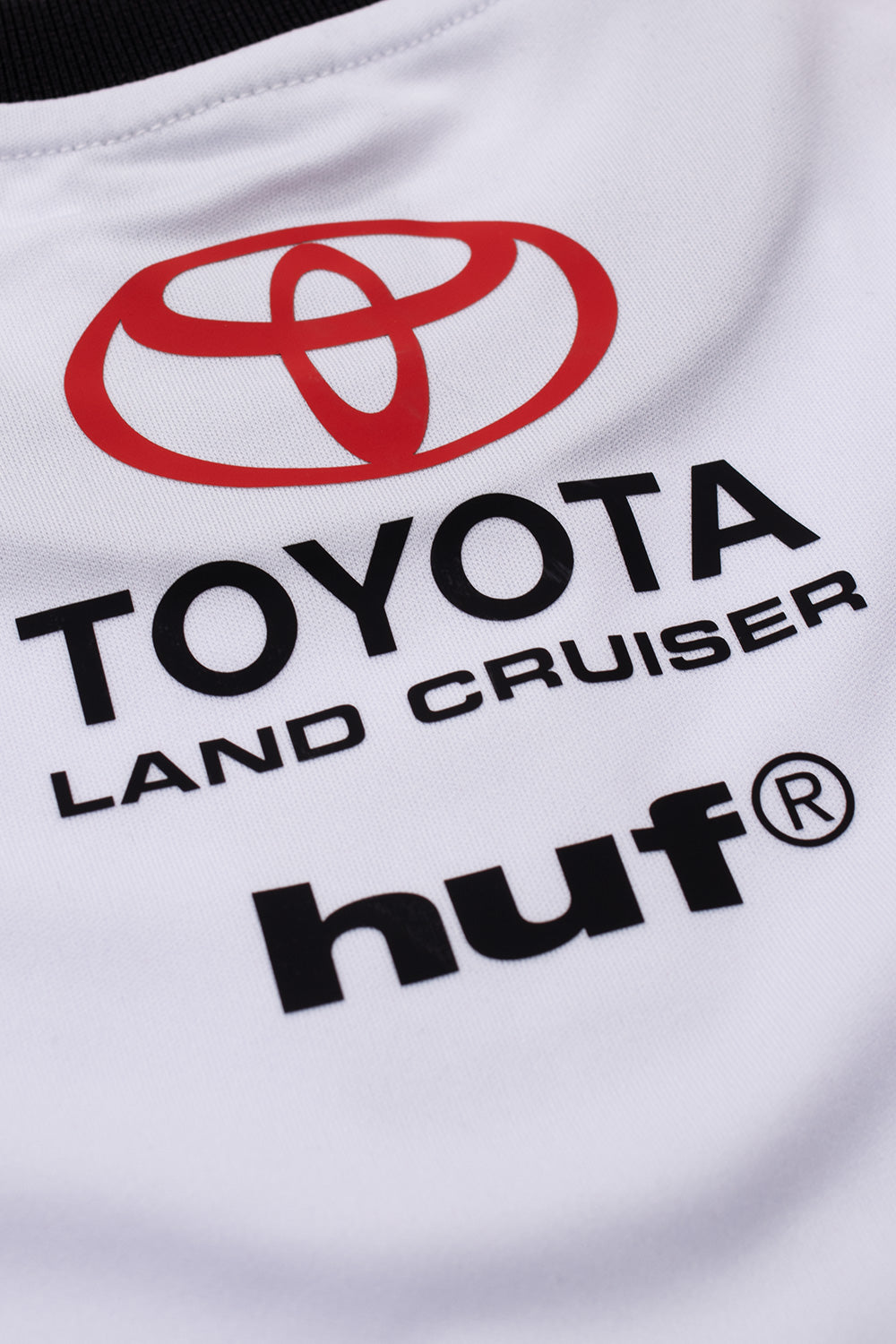 Macro detail of the upper back neckline on the Huf X Toyota Land Cruiser Rally Jersey, focusing on the black finely ribbed collar band and the stacked print consisting of the red Toyota oval badge, "LAND CRUISER" subline, and the bold black "huf®" logo.