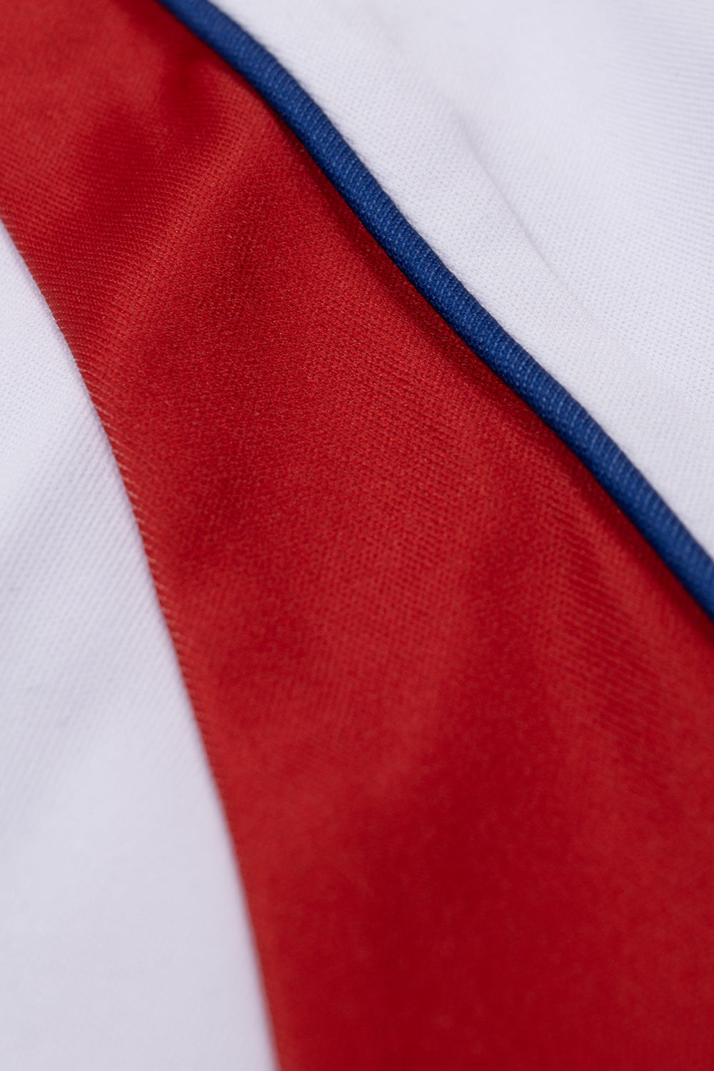 Macro studio texture shot of the raglan shoulder panel on the Huf X Toyota Land Cruiser Rally Jersey, showcasing the smooth technical weave of the red polyester fabric bordered by raised royal blue piping and flatlock seam stitching.