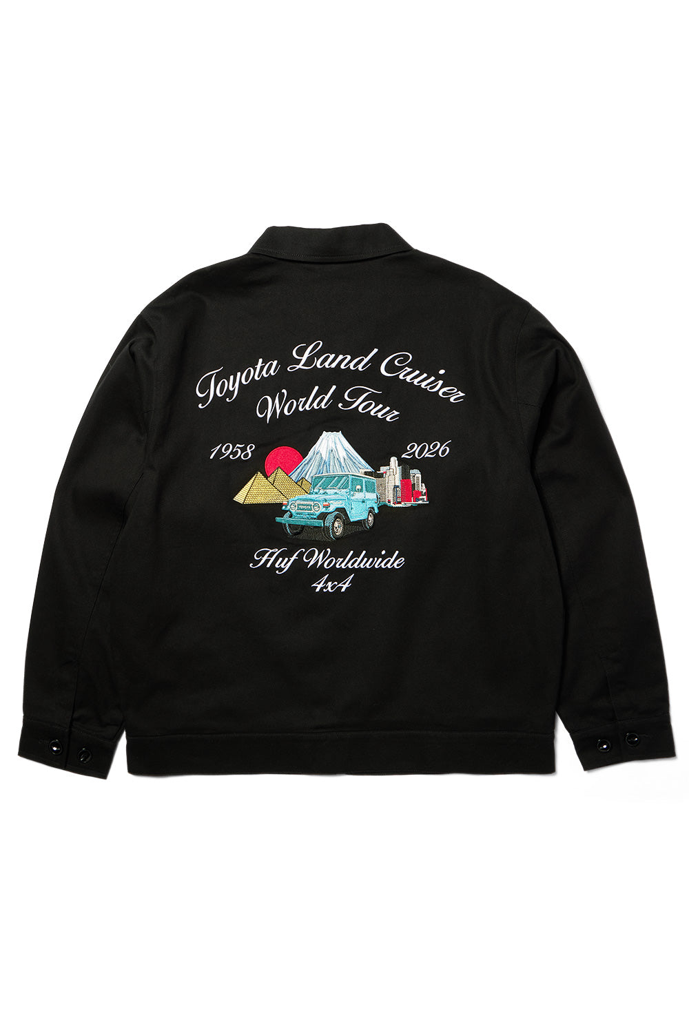 Back flat lay view of the black Huf X Toyota Land Cruiser World Tour Jacket, featuring a wide workwear cut in heavy 280g cotton twill, turn-down collar, button cuffs, and intricate multi-color chainstitch embroidery showing a vintage light blue FJ40 Land Cruiser navigating between the Giza Pyramids, Mount Fuji, and city skyscrapers beneath white scripted lettering reading "Toyota Land Cruiser World Tour 1958-2026" and "Huf Worldwide 4x4"