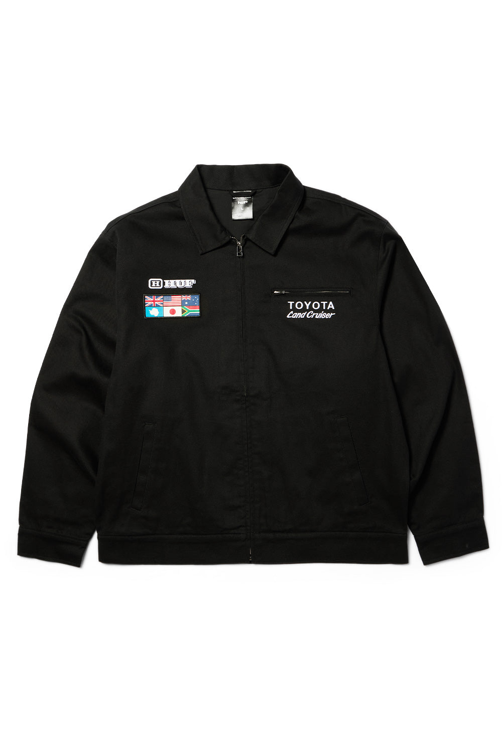 Front flat lay view of the black Huf X Toyota Land Cruiser World Tour Jacket, displaying the full front metal zipper, pointed fold-down collar, angled side welt hand pockets, a zippered horizontal chest pocket with white embroidered "TOYOTA Land Cruiser" lettering, and an embroidered black-and-white HUF logo above a multi-national flag patch on the right chest