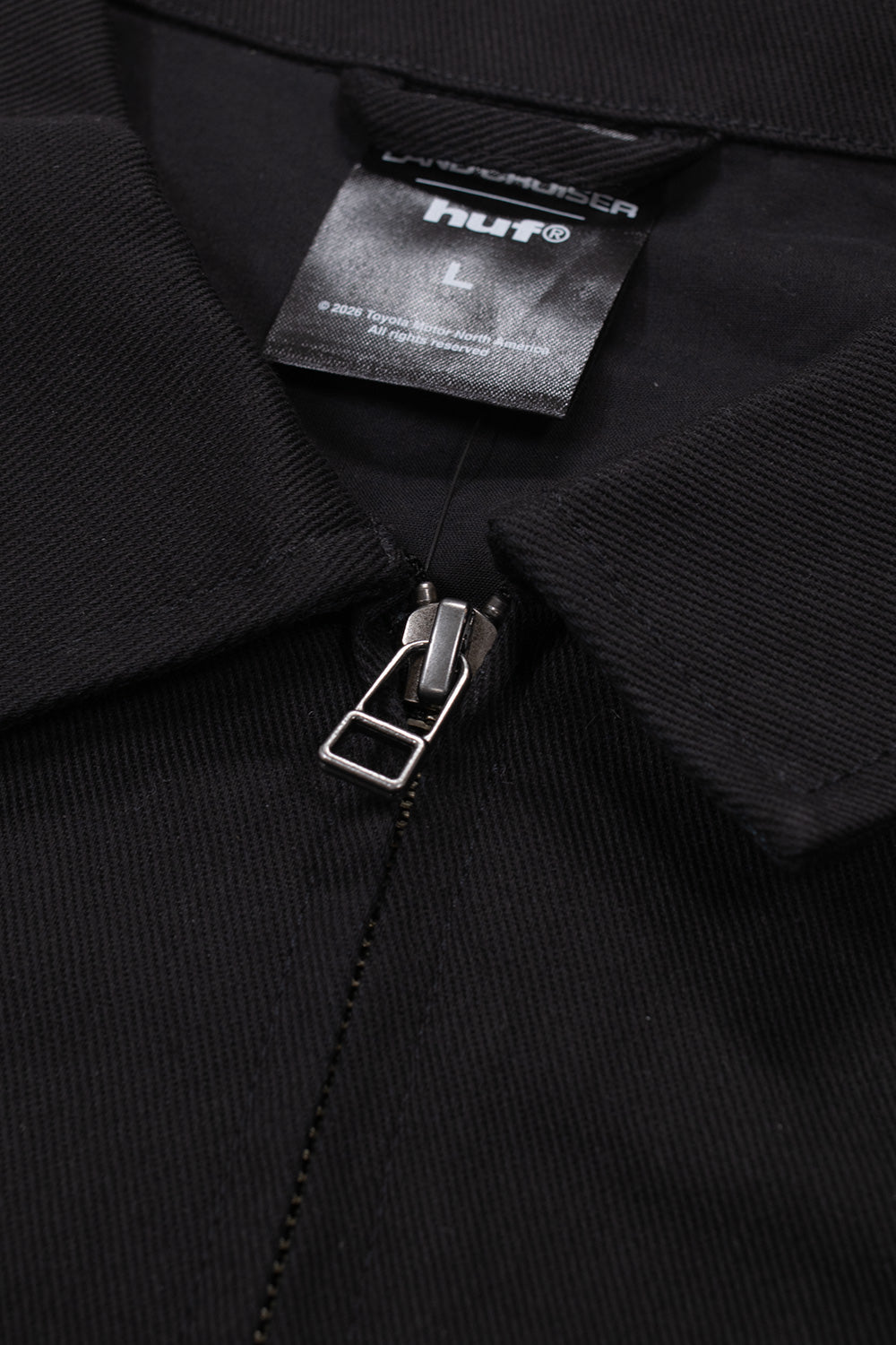 Macro studio close-up of the neckline and collar on the Huf X Toyota Land Cruiser World Tour Jacket, highlighting the silver YKK metal zipper pull, structured black cotton twill lapel, and internal woven black brand collaboration label marked "LAND CRUISER huf® L"