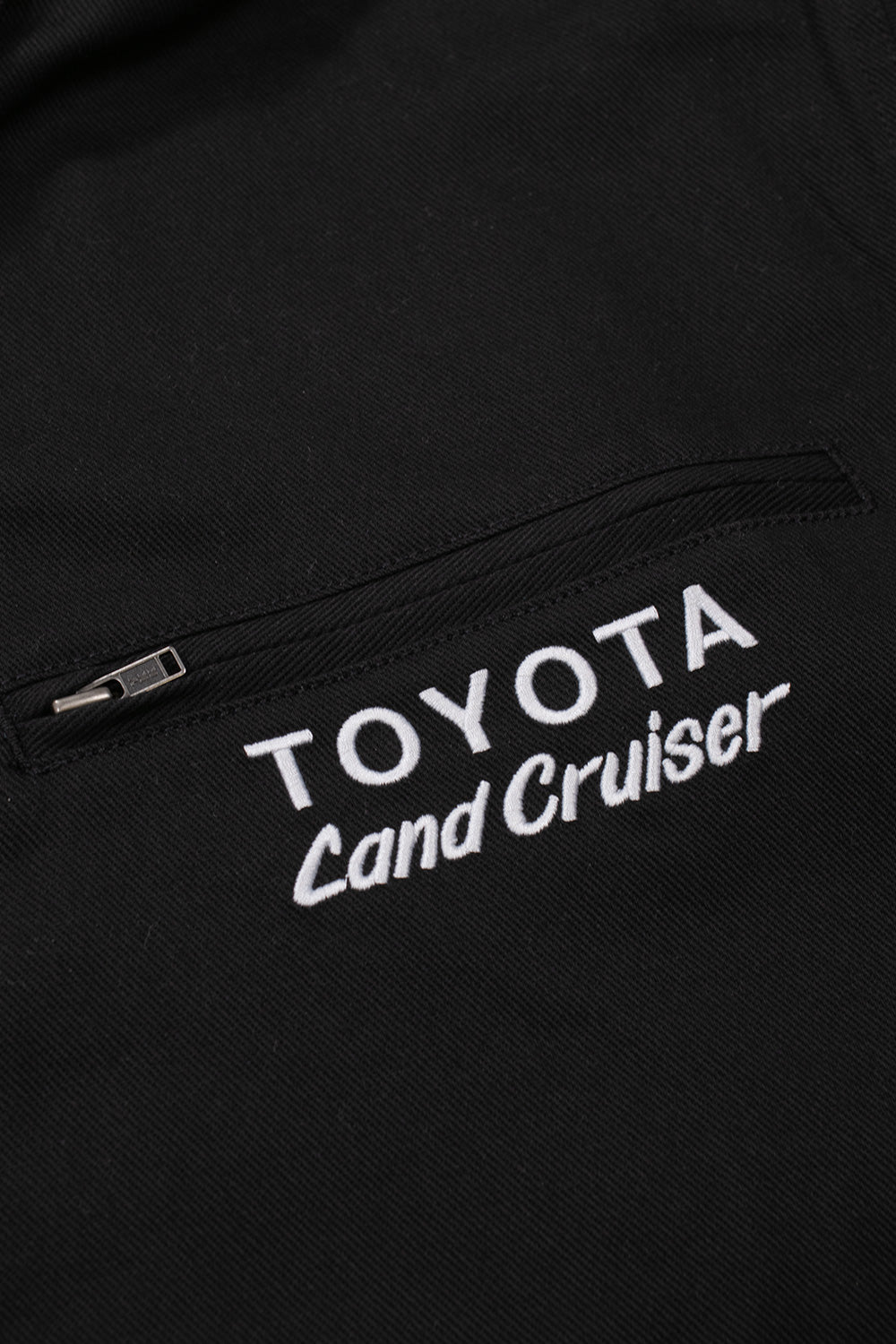 Close-up shot of the left chest pocket on the black Huf X Toyota Land Cruiser World Tour Jacket, showcasing the concealed zipper closure with a small pull tab and clean, dense white satin-stitched embroidery reading "TOYOTA Land Cruiser" on the diagonal twill fabric