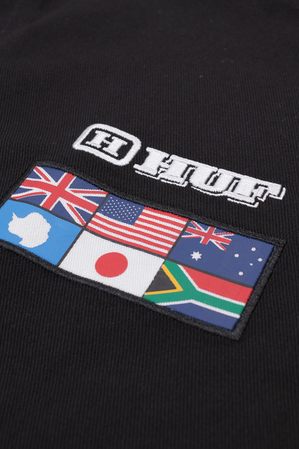 Detailed macro view of the right chest branding on the Huf X Toyota Land Cruiser World Tour Jacket, displaying the bold white outlined embroidered "HUF" block logo positioned directly above a stitched rectangular patch featuring national flags of Antarctica, the United States, Australia, Japan, and South Africa