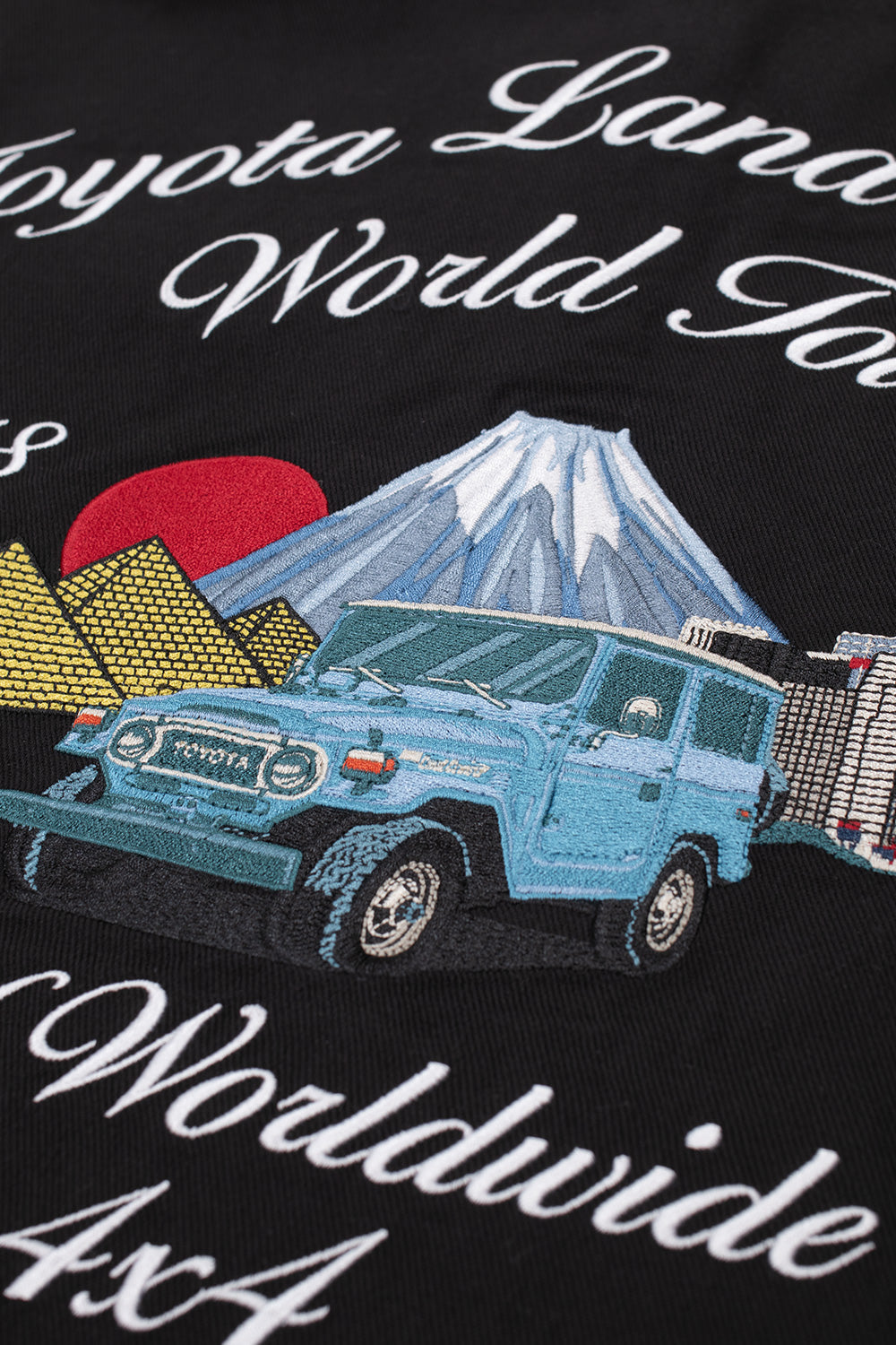Macro studio perspective of the vibrant back embroidery on the Huf X Toyota Land Cruiser World Tour Jacket, highlighting the detailed threadwork of the vintage turquoise Toyota 4x4 vehicle, yellow Egyptian pyramids, red rising sun, snow-capped Mount Fuji, and white script lettering