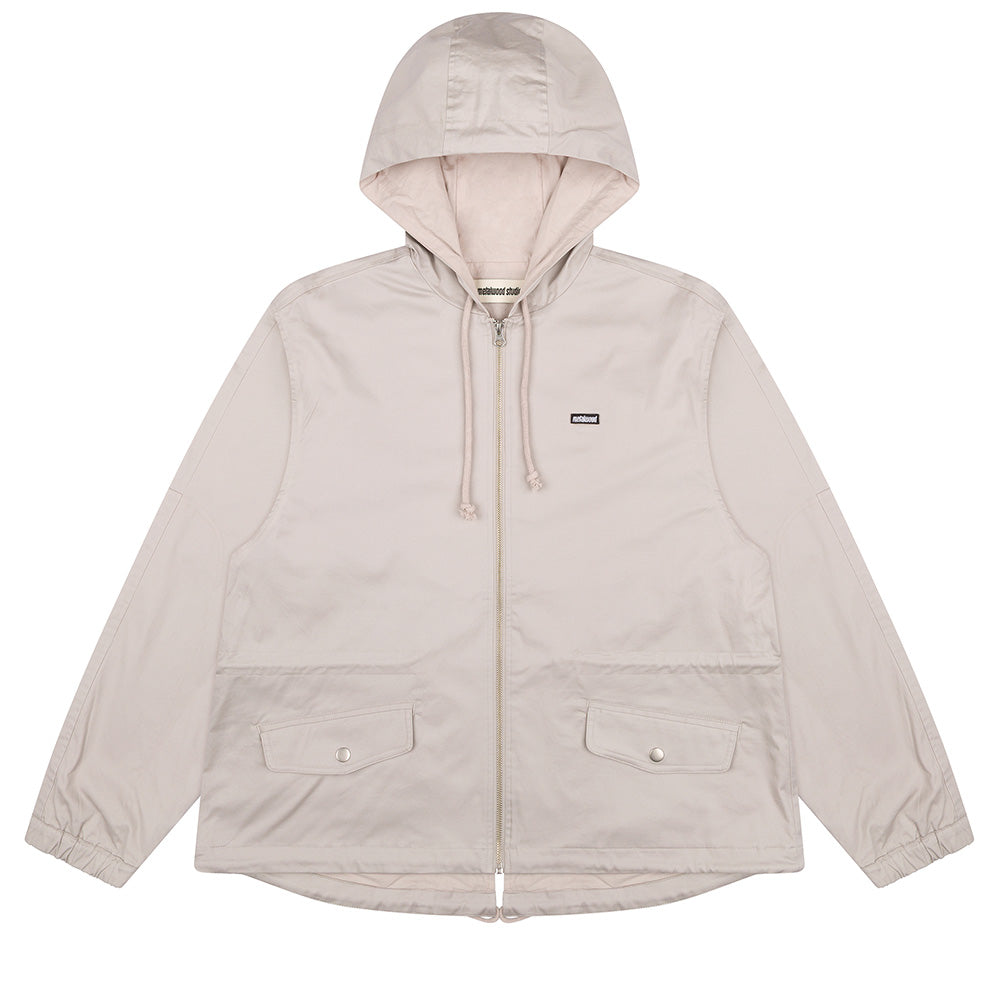 Front flat lay studio view of the Metalwood Studio Cotton Twill Parka in Stone beige, showcasing a lightweight zip-up hooded jacket with tonal rope drawstrings, full-length silver zipper, small black embroidered logo patch on the left chest, two large angled front flap pockets with silver snap closures, elasticated wrist cuffs, and an adjustable curved drawstring hem.