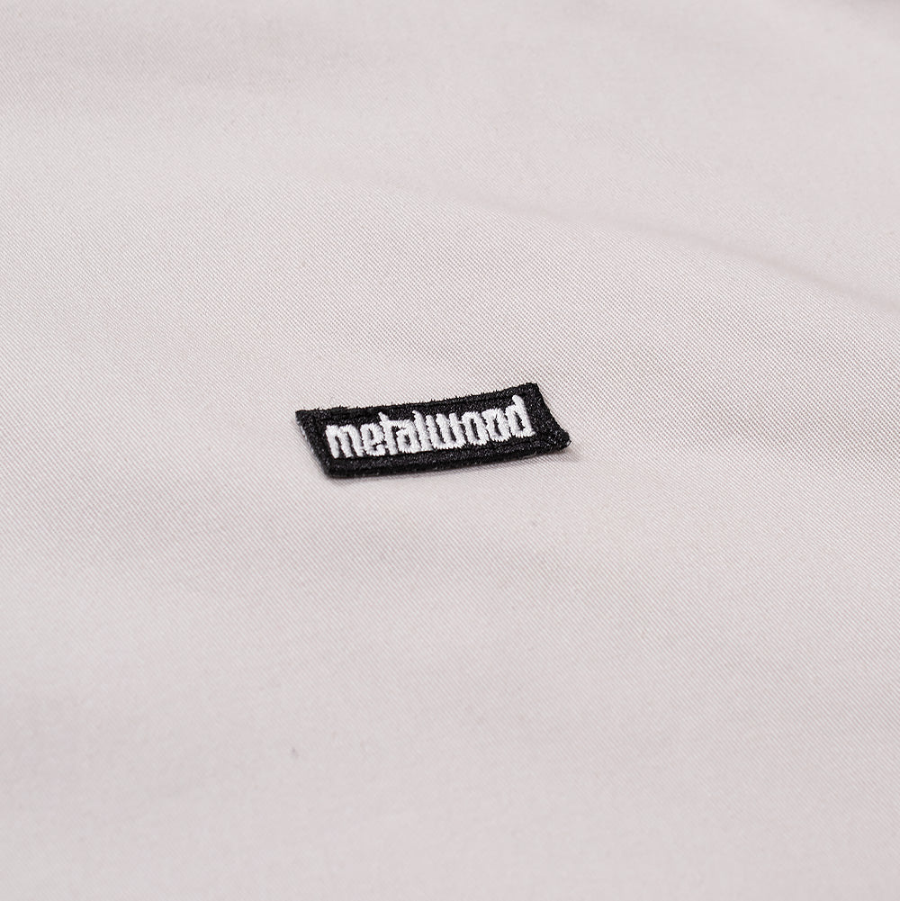 Detailed macro perspective of the chest branding on the Metalwood Studio Cotton Twill Parka, highlighting the small black rectangular woven patch embroidered with contrasting white lowercase "metalwood" typography stitched onto smooth stone-beige cotton twill.