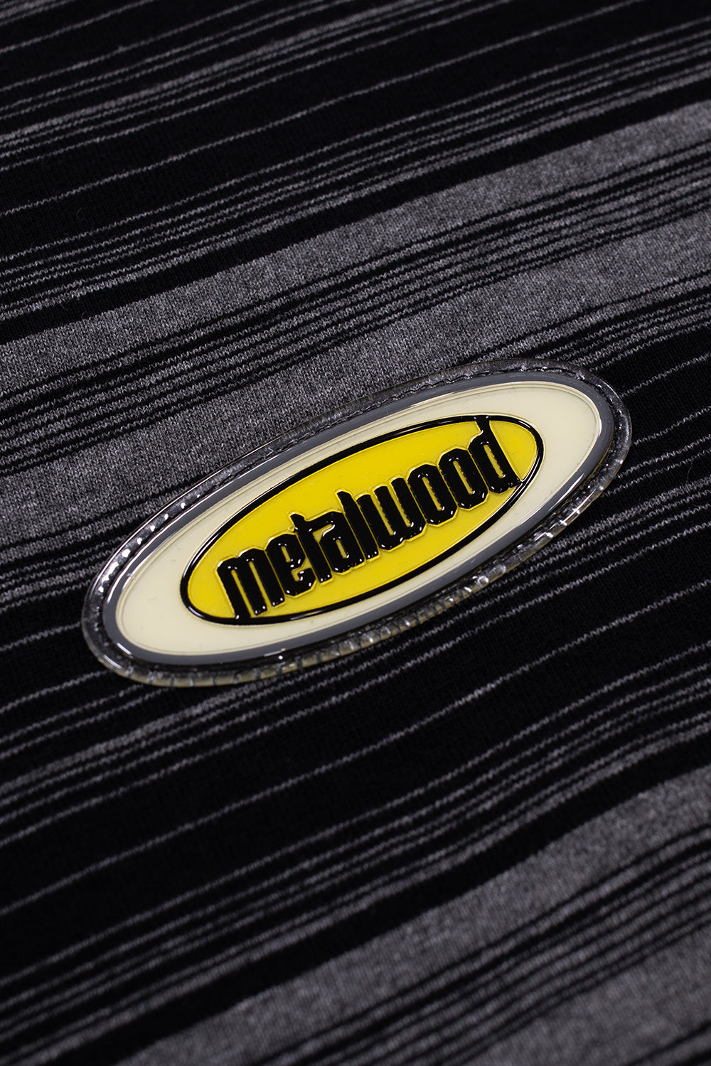 Close-up of Metalwood Studio rubber chest patch on black striped cotton T-shirt, showcasing premium branding, textured fabric construction and retro golf-meets-skate streetwear detailing