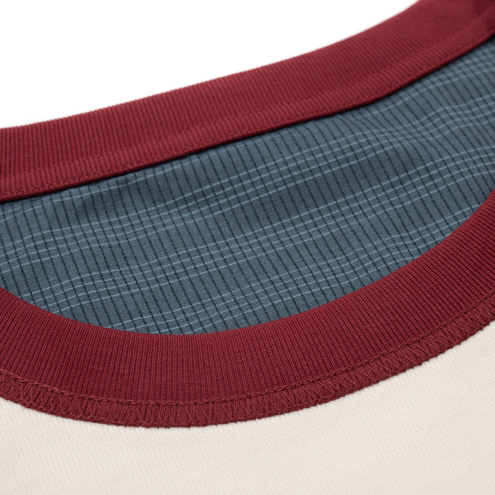 Macro close-up perspective of the neckline on the Metalwood Studio Dual Face T-Shirt, highlighting the dense burgundy rib-knit collar trim with reinforced overlock stitching set against the smooth bone-white cotton outer face and revealing the inner blue-grey technical grid-patterned lining.