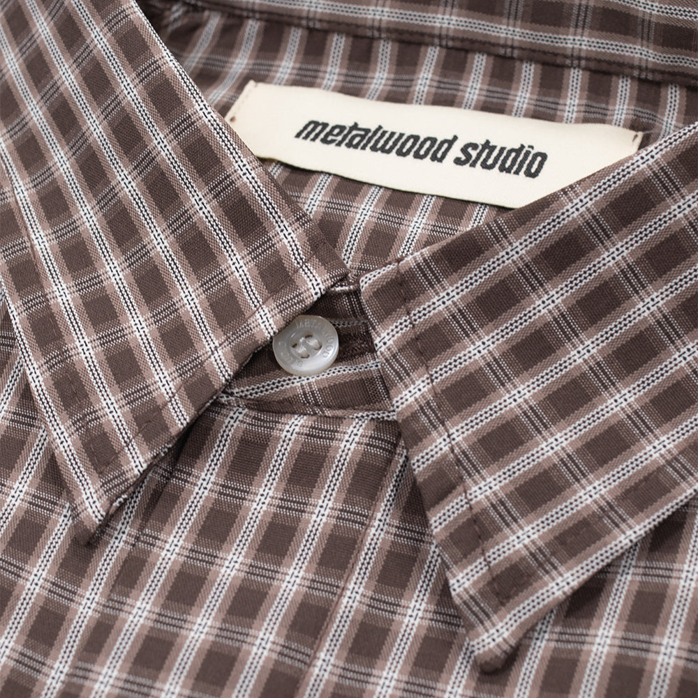 Macro studio close-up of the collar and neckline on the Metalwood Studio Frankenstein Shirt, detailing the pointed dress collar in brown check twill, engraved white pearlized top collar button, and the woven cream "metalwood studio" brand label along the inner neckband.