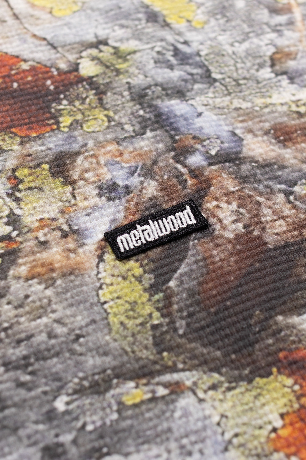 Close-up of the sewn-on Metalwood Studio mini logo patch on the Real Moss Camo Thermal Shirt, highlighting textured waffle knit cotton fabric and detailed camo print.