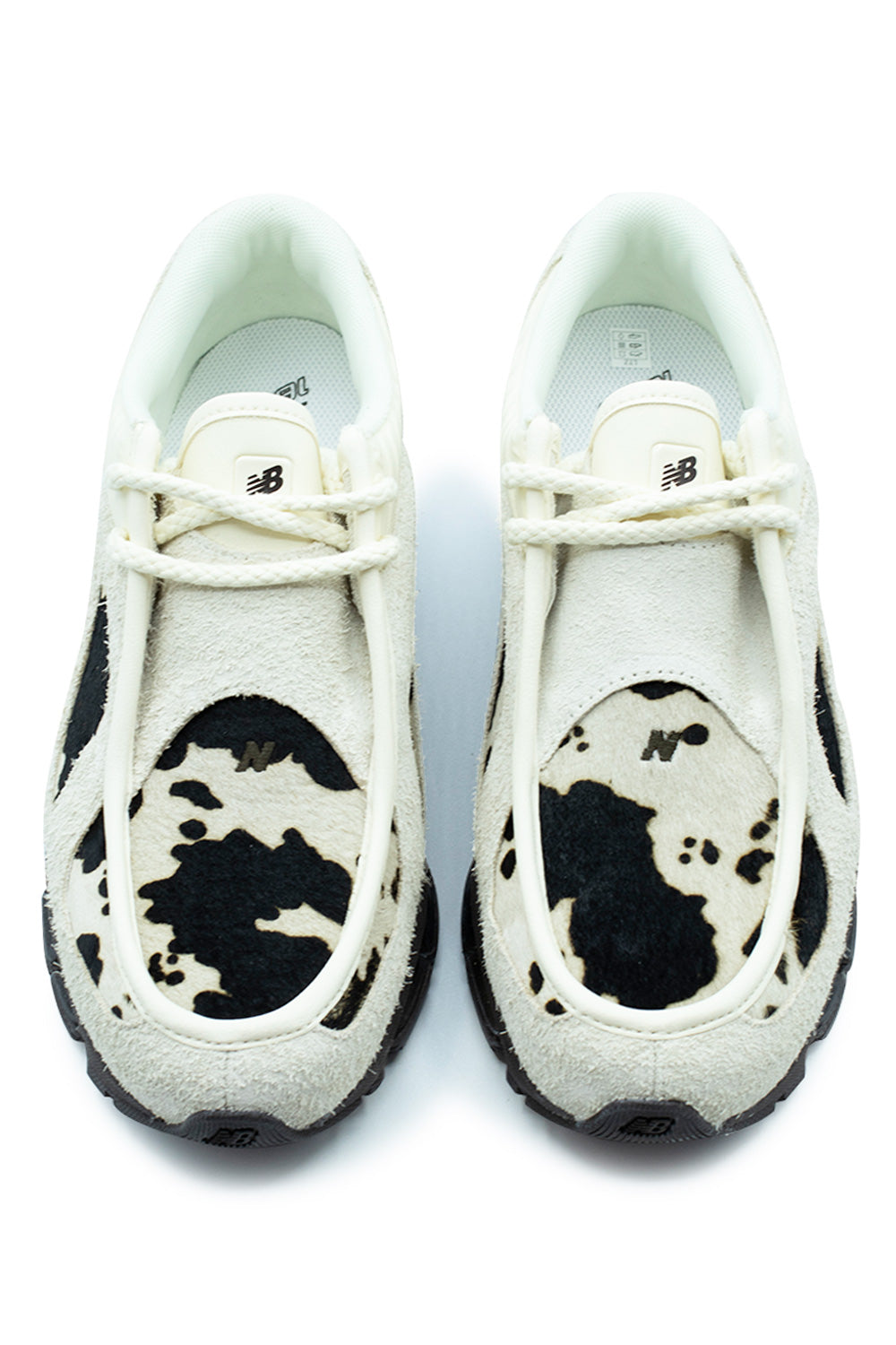 Top-down studio shot of a pair of New Balance 1890A shoes, highlighting the tactile cow-pattern pony hair toe vamp panels accented by subtle tonal "N" logos, raised piped moccasin apron seam border in hairy Angora suede, thick round cream cord laces, and short padded leather tongues with black "NB" branding.