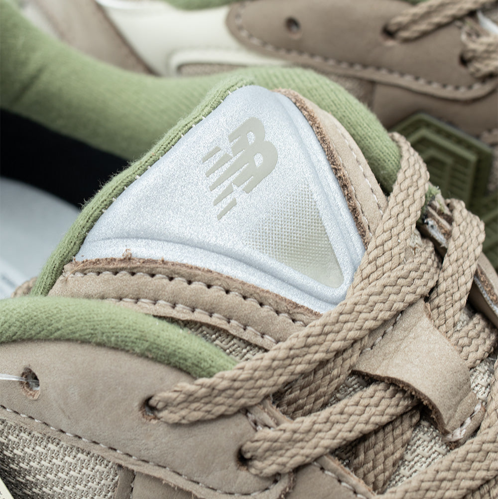 Macro studio close-up of the tongue and lacing system on the New Balance 2010 GORE-TEX in Mushroom, capturing the reflective silver triangular tongue badge with embossed NB branding, thick woven beige oval laces, soft olive green collar padding, and detailed double-stitched tan nubuck eyestays.