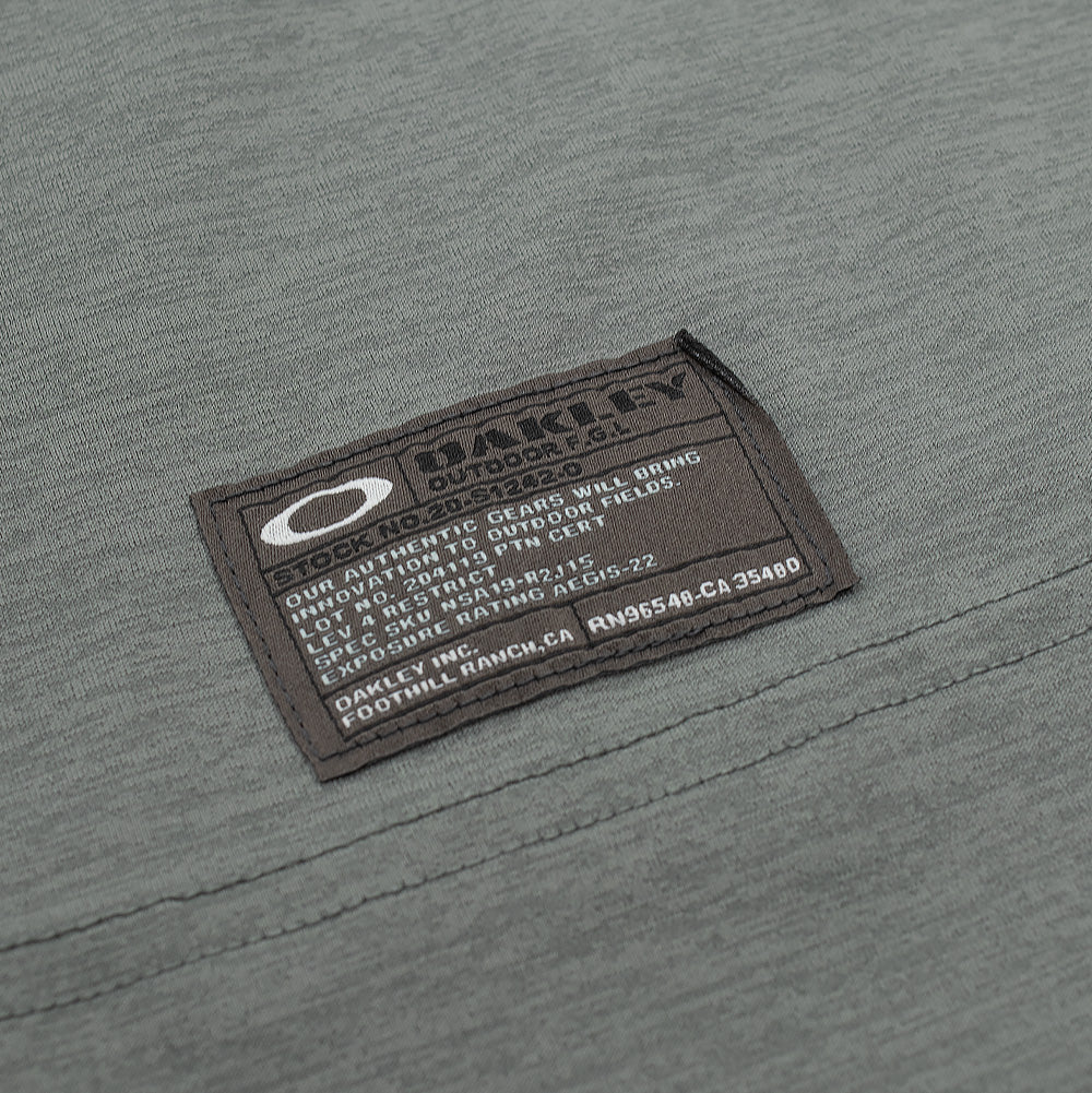 Macro studio close-up of the lower hem label on the Oakley FGL Scratch SS Tee 6.7 in Acrid Green, displaying the rectangular dark woven field spec patch with the white Oakley Ellipse icon, "OAKLEY OUTDOOR F.G.L." lettering, and authentic product codes stitched neatly above the hem seam.