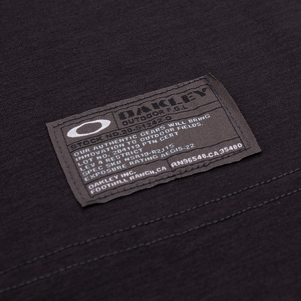 Macro studio close-up of the hem label on the Oakley FGL Scratch SS Tee 6.7 in Phantom, displaying the rectangular woven field specification patch featuring the white Oakley Ellipse icon, "OAKLEY OUTDOOR F.G.L." insignia, and tactical serial rating text.