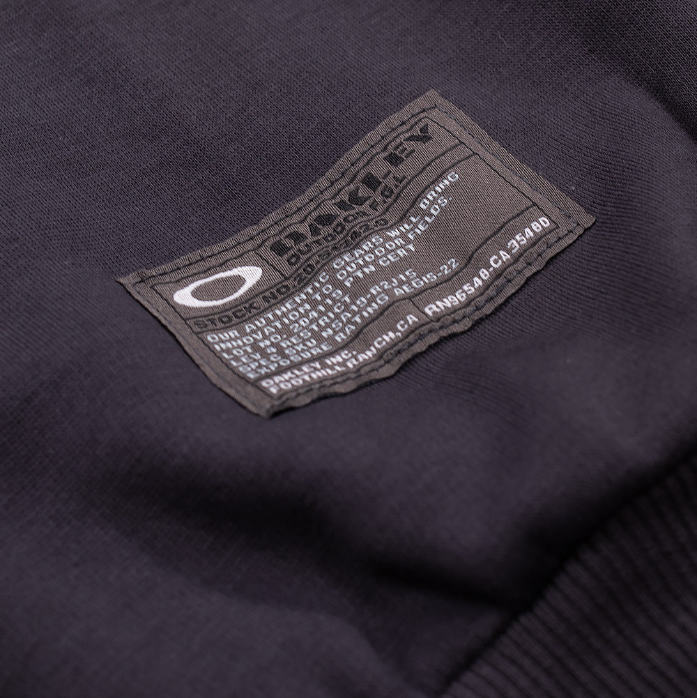 Macro studio close-up of the left sleeve cuff branding on the Oakley FGL Slick Fleece Hoodie 6.7 in Phantom, showing the rectangular military-style woven label with white Oakley Ellipse branding, "OAKLEY OUTDOOR F.G.L." insignia, and technical specification codes stitched directly above the ribbed wrist trim.