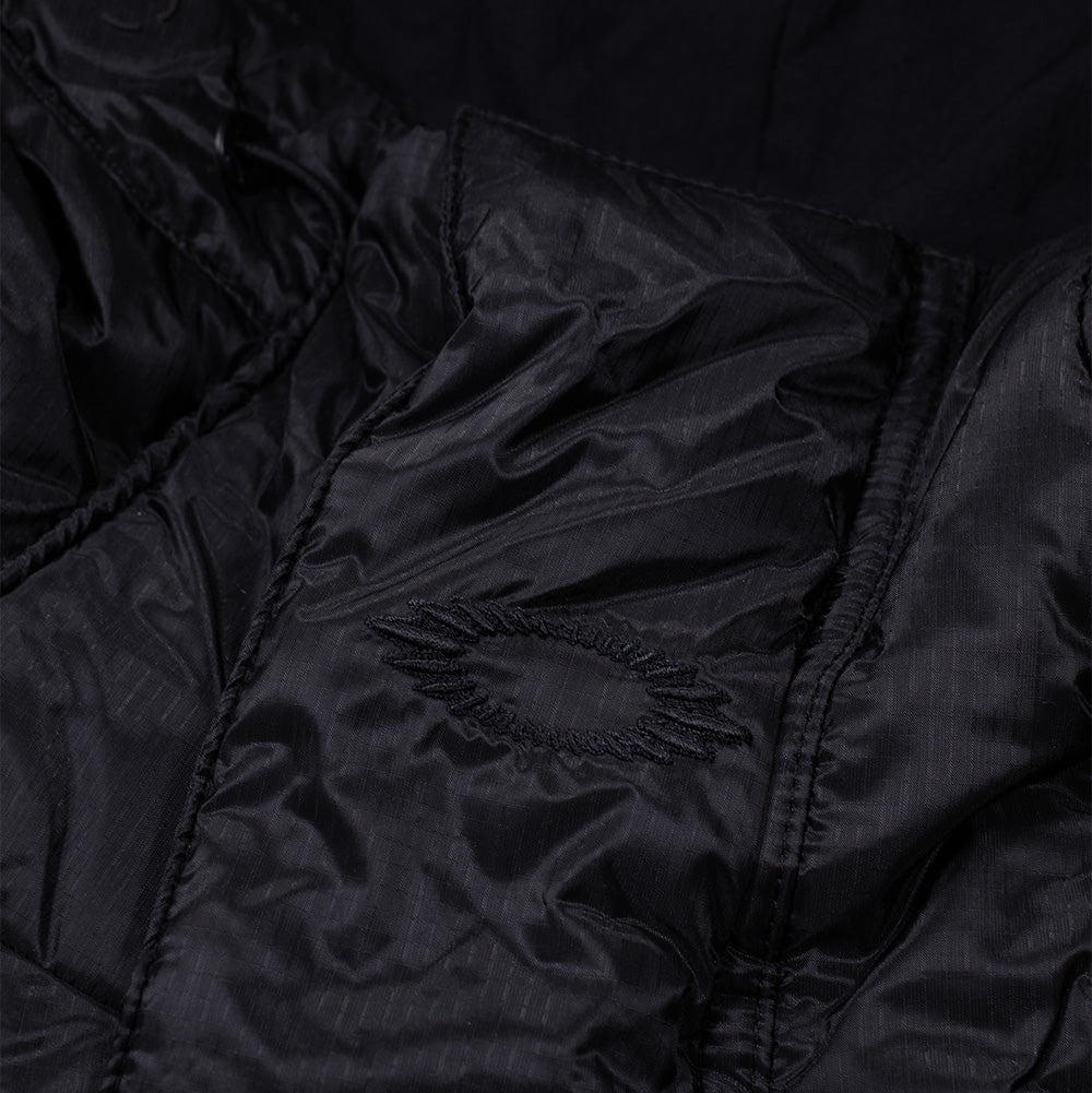 Detailed macro perspective of the upper chest branding on the Oakley FGL Stealth Ins Jacket 2.0, focusing on the tonal black satin-stitch embroidery forming the jagged, radiating scratch-style Oakley Ellipse emblem set into the textured micro-ripstop Schoeller shell.